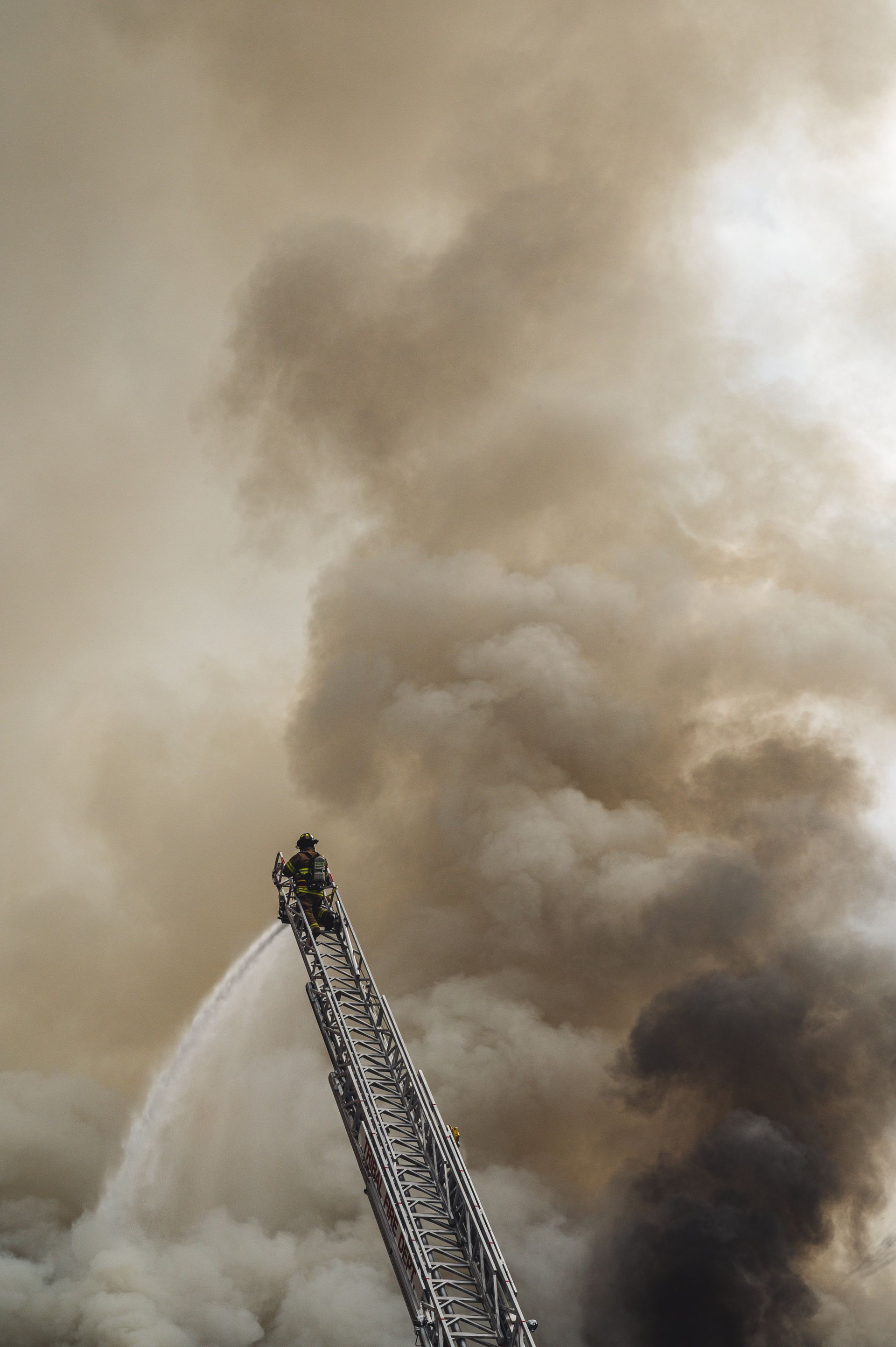 A firefighter on a ladder spraying water on fire with lots of smoke