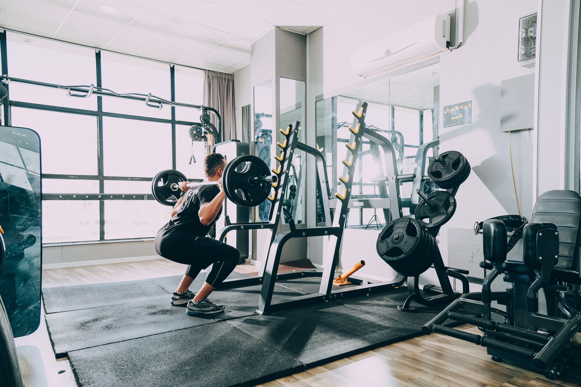 A well-lit residential fitness space, showing various fitness equipment and a person performing the squat lift with weight on the barbell.
