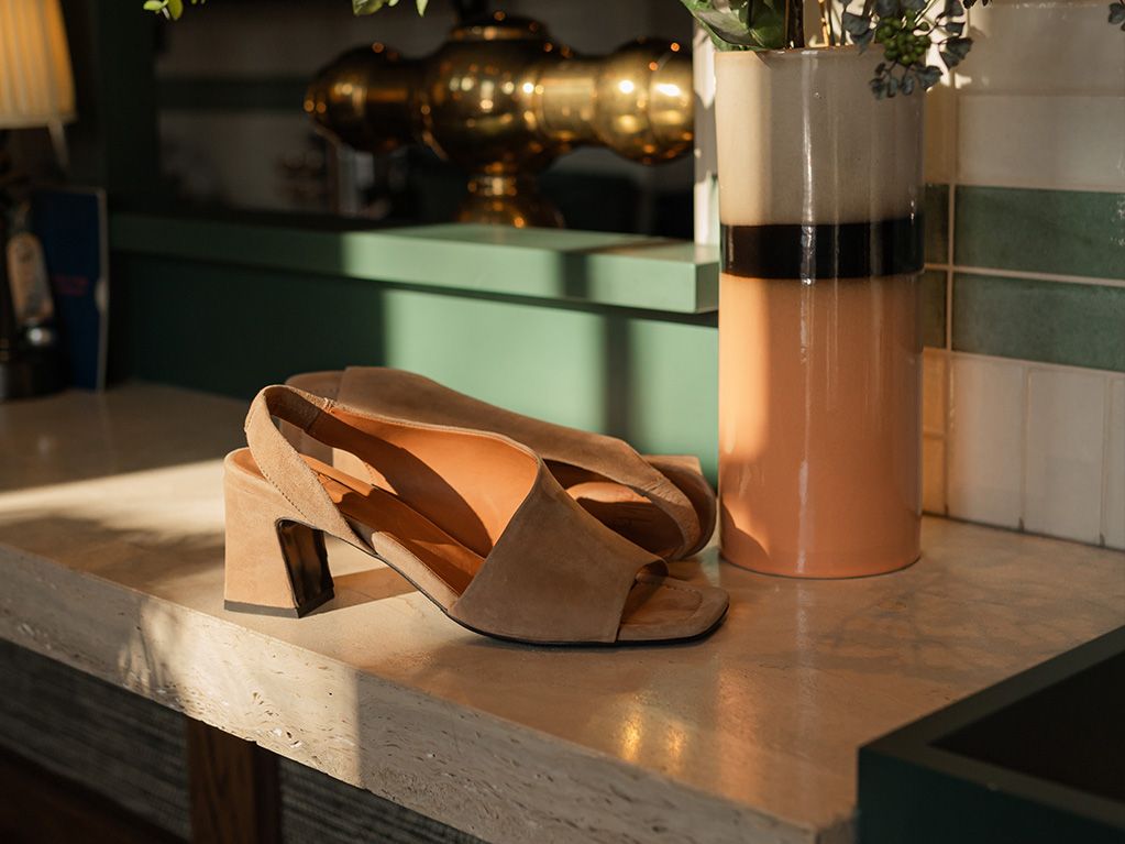 Tan suede high-heeled sandals with open toes placed on a marble surface near a decorative ceramic vase in warm sunlight.