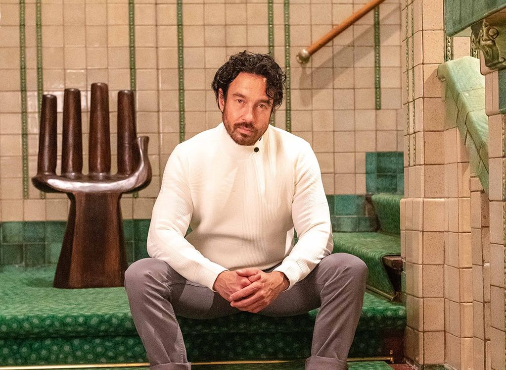 Man zittend op groen beklede trap met Koll3kt off-white sweater met knoopjes