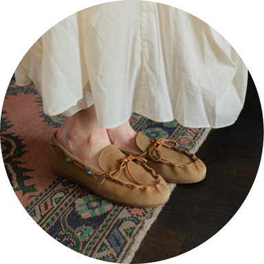 Person wearing tan moccasins with turquoise beads, standing on a patterned rug. The edge of a white flowing skirt is visible.