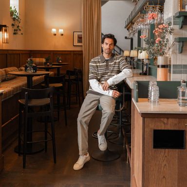 Man in a striped sweater and beige pants sits on a stool in a cozy café with wooden decor and floral accents.