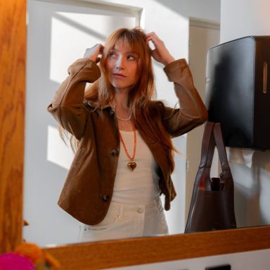 Woman in a brown jacket adjusts her hair while looking in a mirror, with sunlight streaming through a window, casting shadows on the wall.