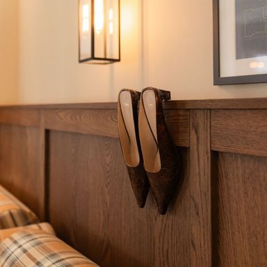 Brown shoes hanging from a wooden wall paneling in a cozy room, beneath soft lighting and near a plaid-patterned upholstered bench.