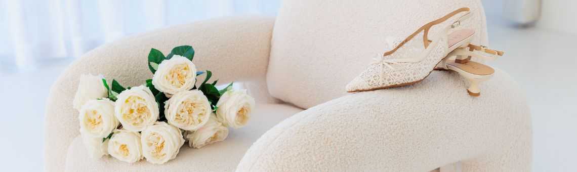 Een paar elegante kanten bruidsschoenen en een boeket witte rozen liggen op een zachte crèmekleurige stoel.