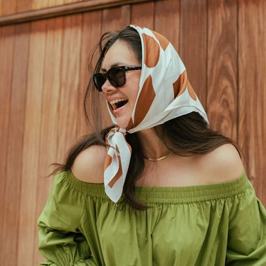 Smiling woman in sunglasses and a patterned headscarf, wearing an off-shoulder green top, standing against a wooden background.