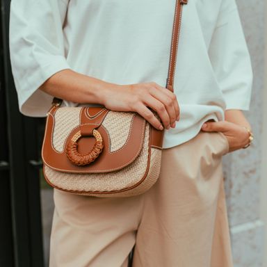 Person wearing a white top and beige pants, holding a stylish woven handbag with brown leather accents and a circular handle.