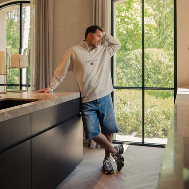 Man in casual attire leans on a kitchen counter, gazing out a window with lush greenery outside.