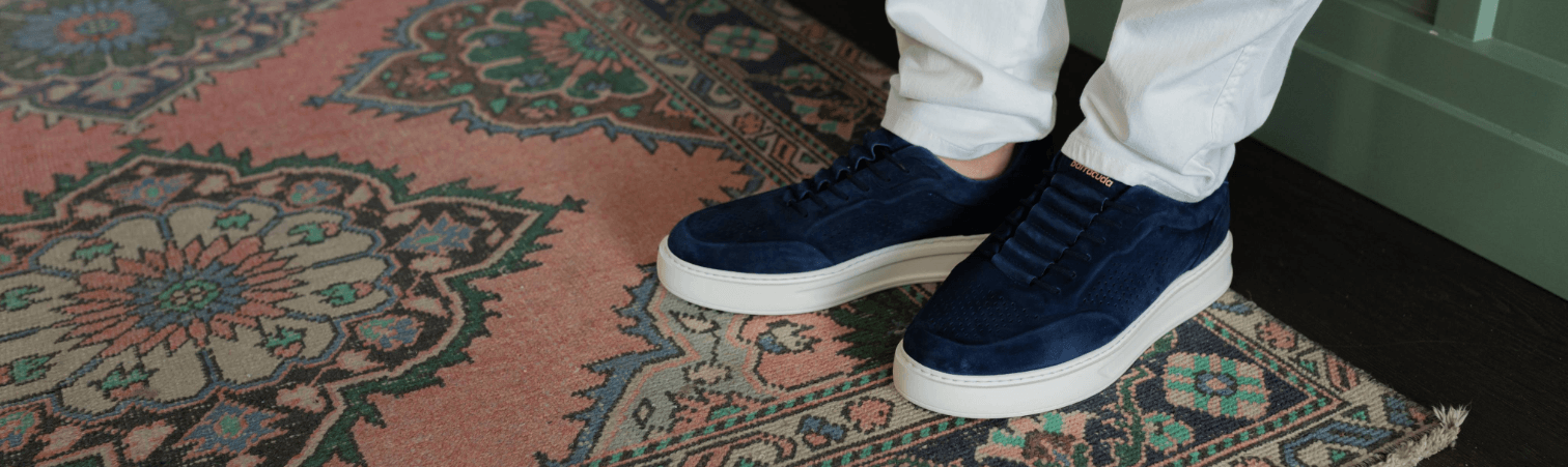 Iemand met blauwe suède sneakers en een witte broek, staand op een kleurrijk vloerkleed met patroon