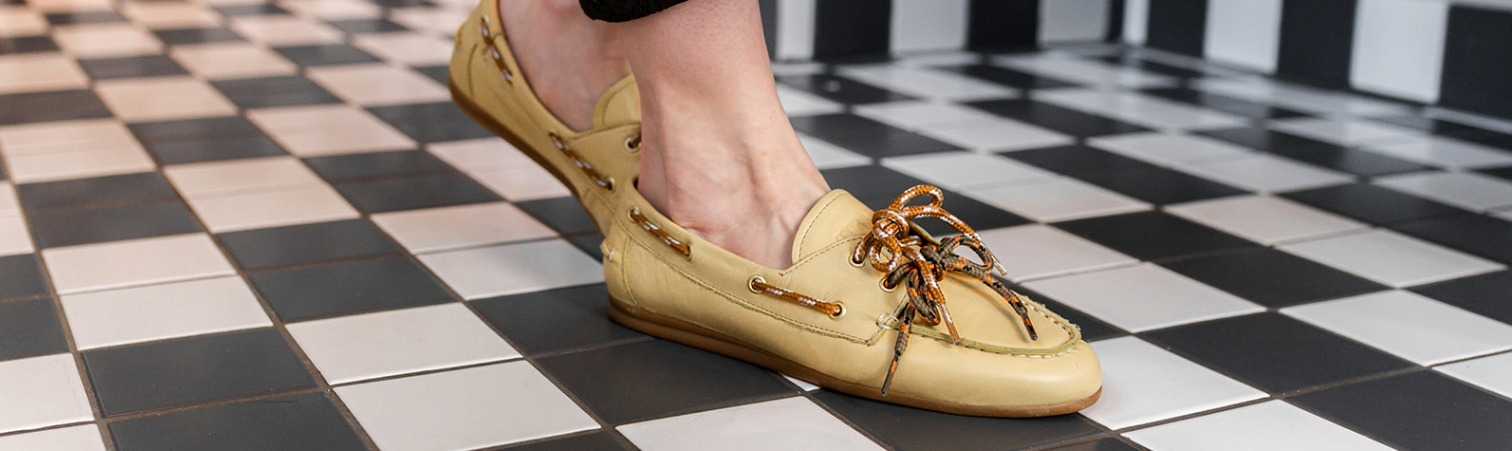 Een persoon die lichtgele leren bootschoenen met decoratieve veters draagt, staat op een zwart-wit geblokte vloer