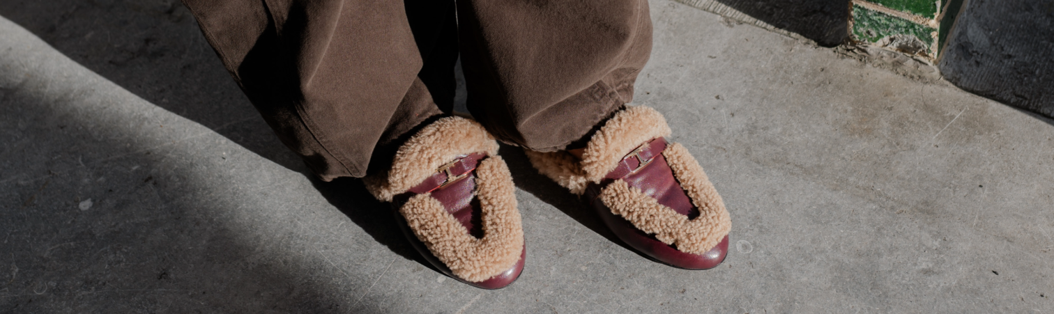 Bovenaanzicht bordeauxrode leren instaploafers met cognackleurige bontrand onder wijdvallende bruine broek