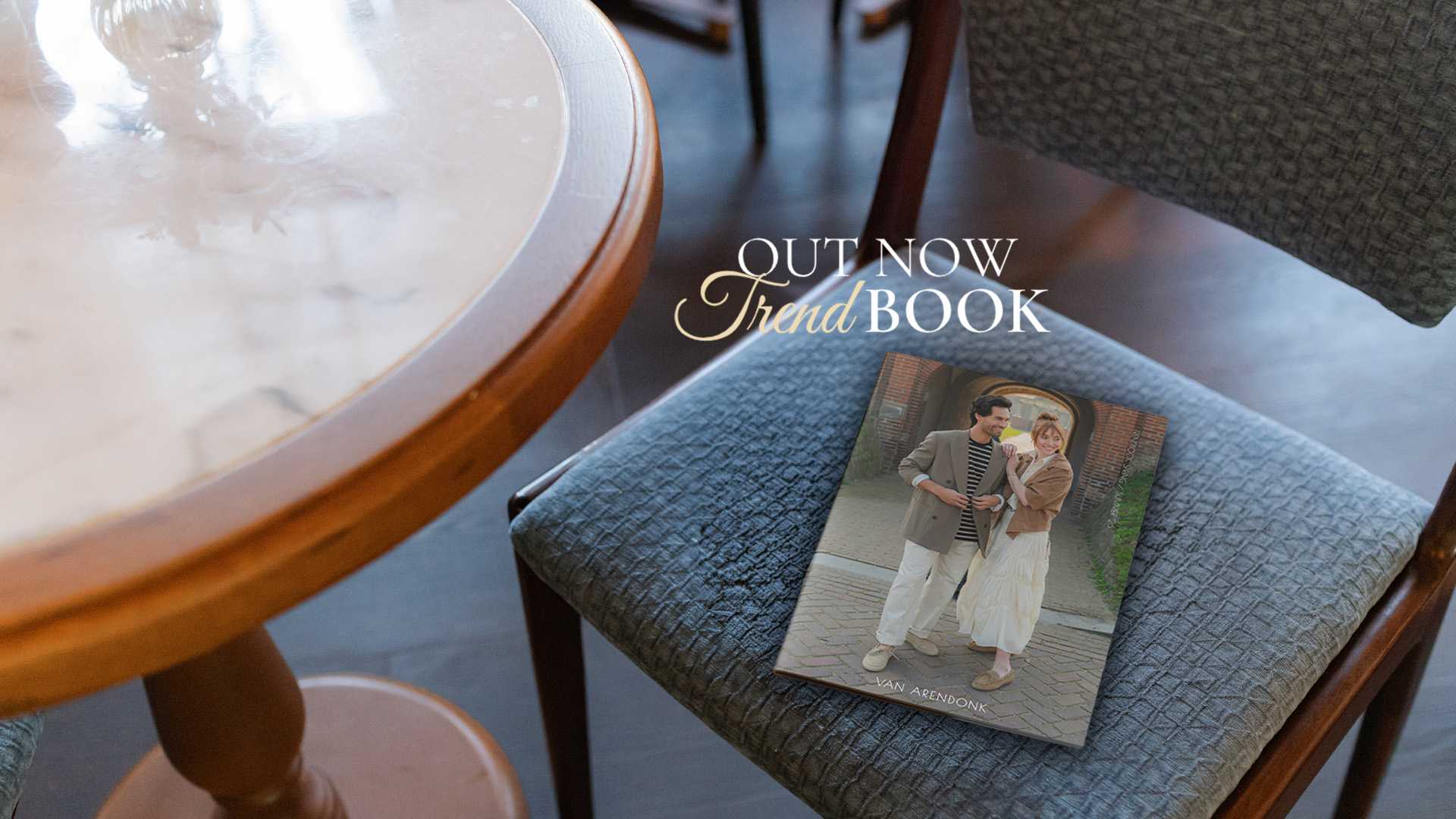 A magazine titled "Out Now Trend Book" lies on a chair next to a round wooden table.