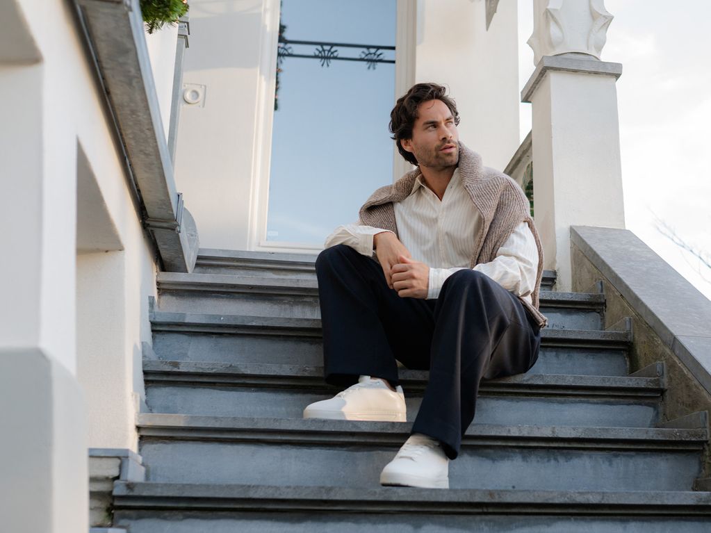 Man sitting on outdoor steps, wearing a beige shirt, dark pants, and white shoes, with a jacket draped over his shoulders.