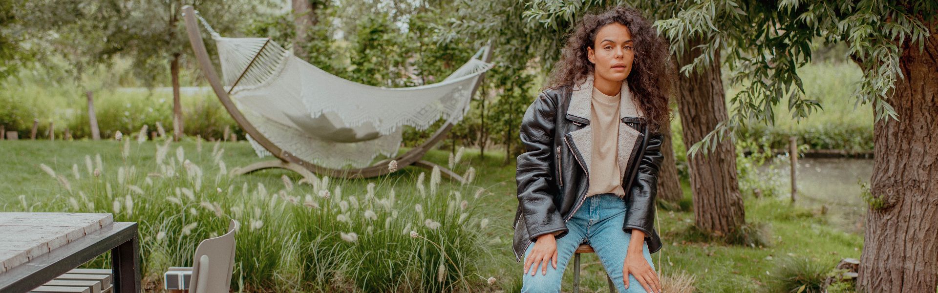 Vrouw met krullend haar en leren lammy jack Goosecraft leunend op krukje in tuin