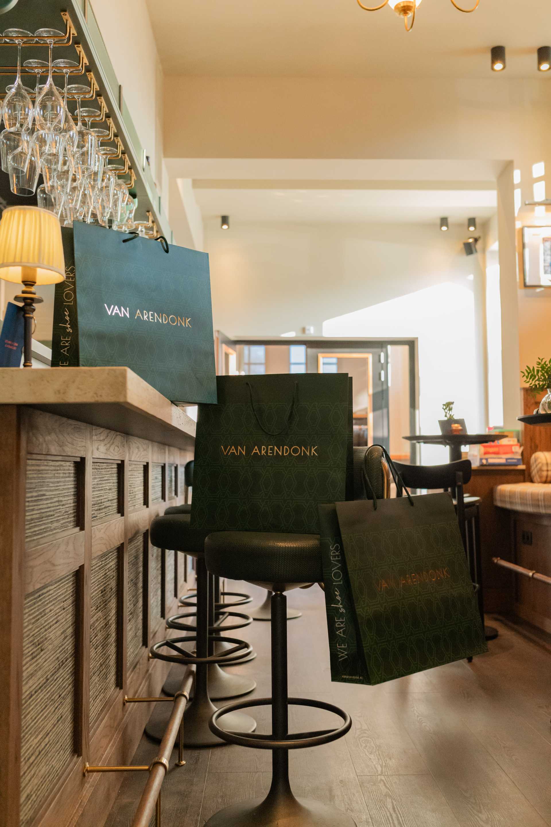 Elegante Van Arendonk tassen op barkrukken in een stijlvol, warm verlicht interieur met hangende glazen en sfeervolle decoratie