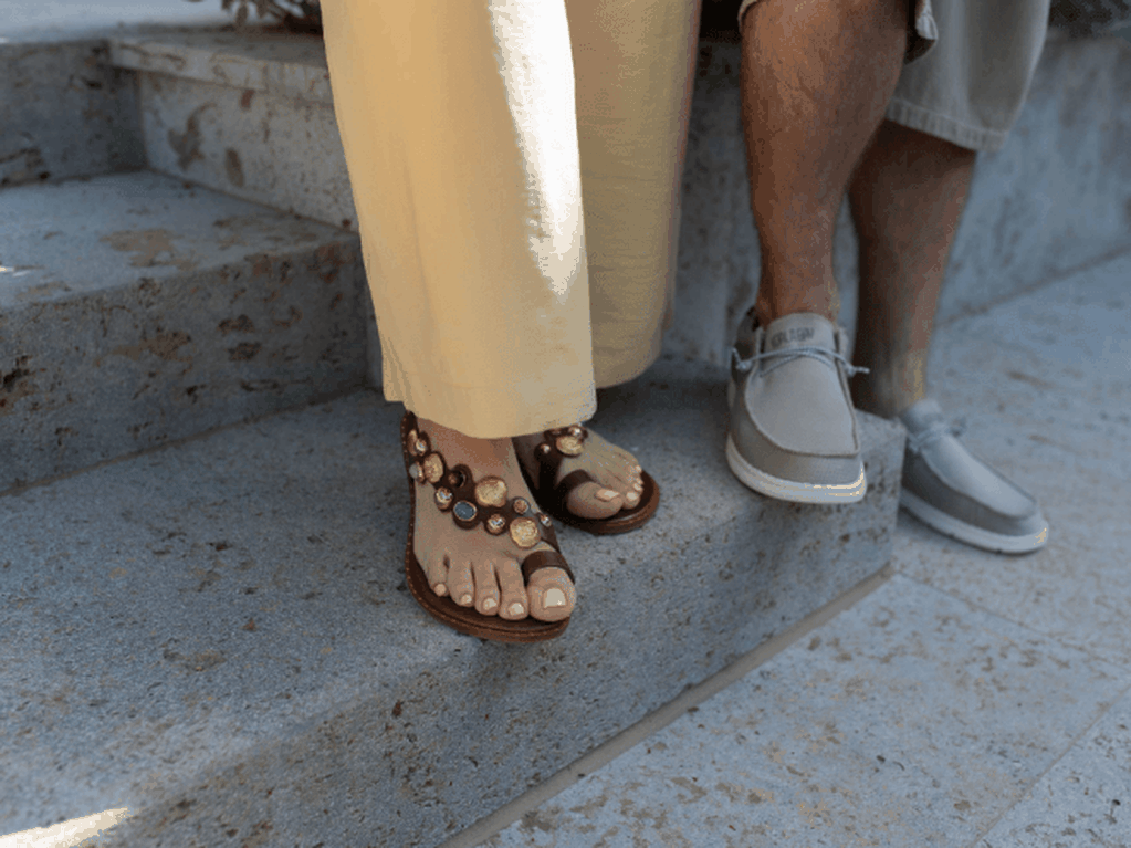 Vrouw met lichtgele broek en bruine sandalen met gekleurde stenen naast man met grijze sportieve bootschoenen op stenen trap