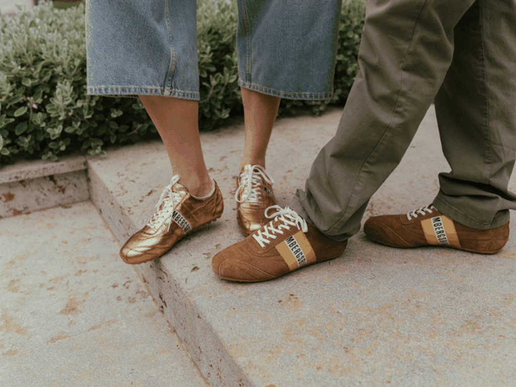 Twee mensen met skinny sneakers aan — de een goud, de ander bruin met gele strepen — staand op een betonnen rand naast het groen