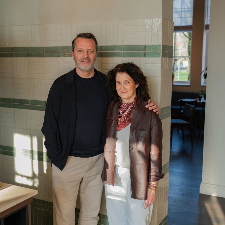 David en Danielle van Arendonk staan samen in een warm verlichte kamer en glimlachen naar de camera, met op de achtergrond betegelde wanden en een raam
