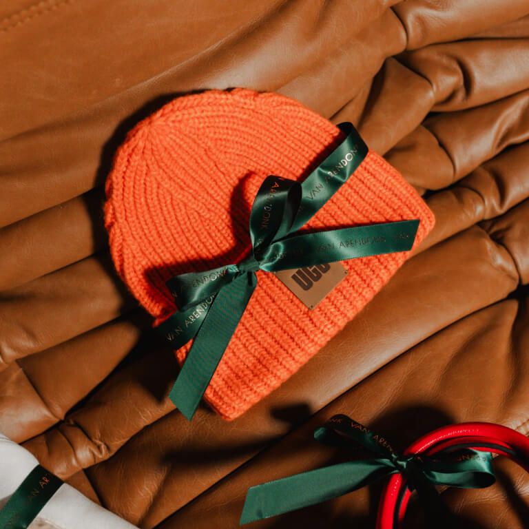 Gebreide oranje UGG beanie met groen strik op cognackleurige leren bank