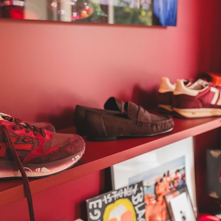 Donkerrode-sneakers-en-bruine-loafers-op-rode-plank-met-kunstwerken-en-fotolijsten-op-de-achtergrond