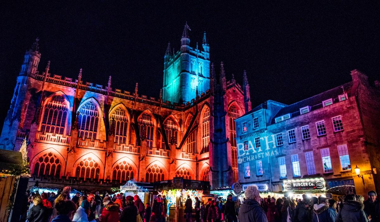 Image of Bath Cathedral at night during a Christmas market, it is illuminated in multi-coloured light