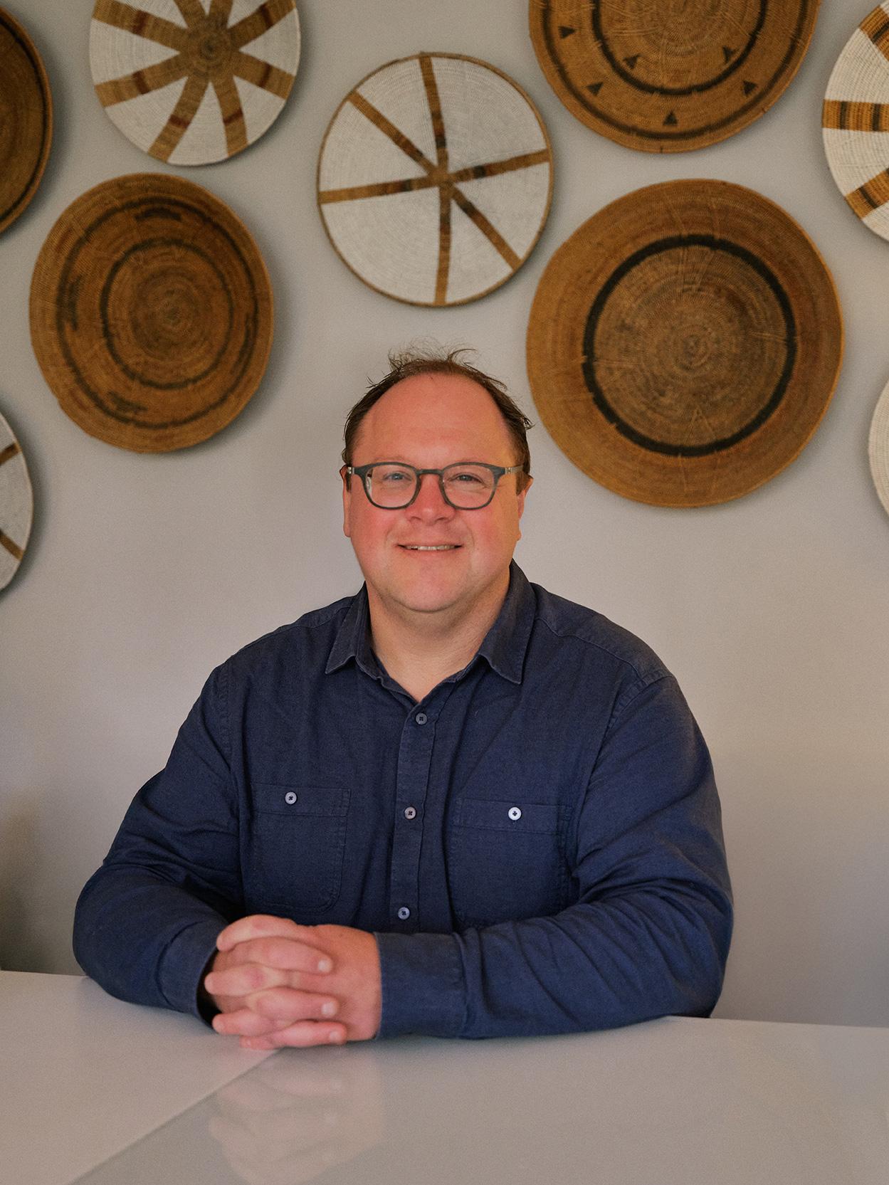 Image of Russell Hunt, Founder of Pets Lets. He is seated at a white table, with circular woven decorations on the wall behind him.