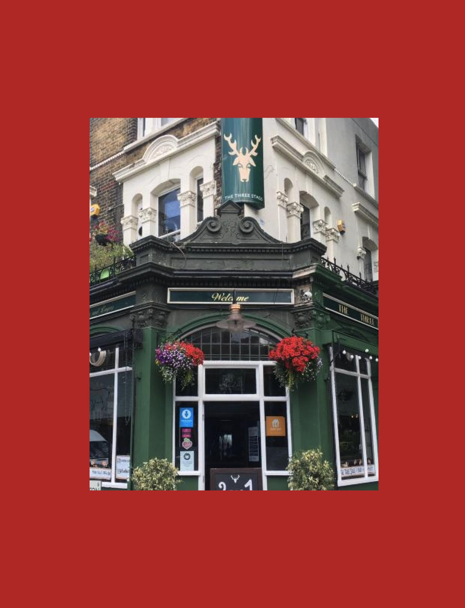 Traditional British pub on a corner, with vibrant flower baskets and '2 for 1 Cocktails' offer. The green facade stands out under a clear sky, inviting guests to its outdoor seating area.