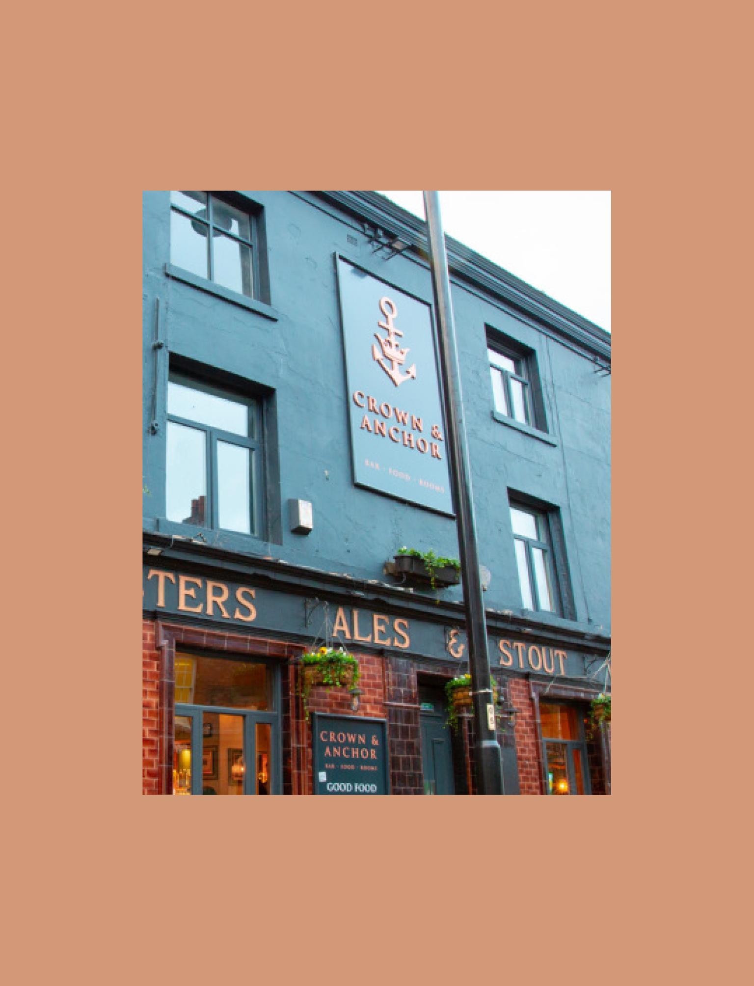 A corner pub painted blue - with a sign saying "Crown &amp; Anchor" on the facing side. It has a banner wrapping around it, listing Ales, Stout, Wines &amp; Spirits.