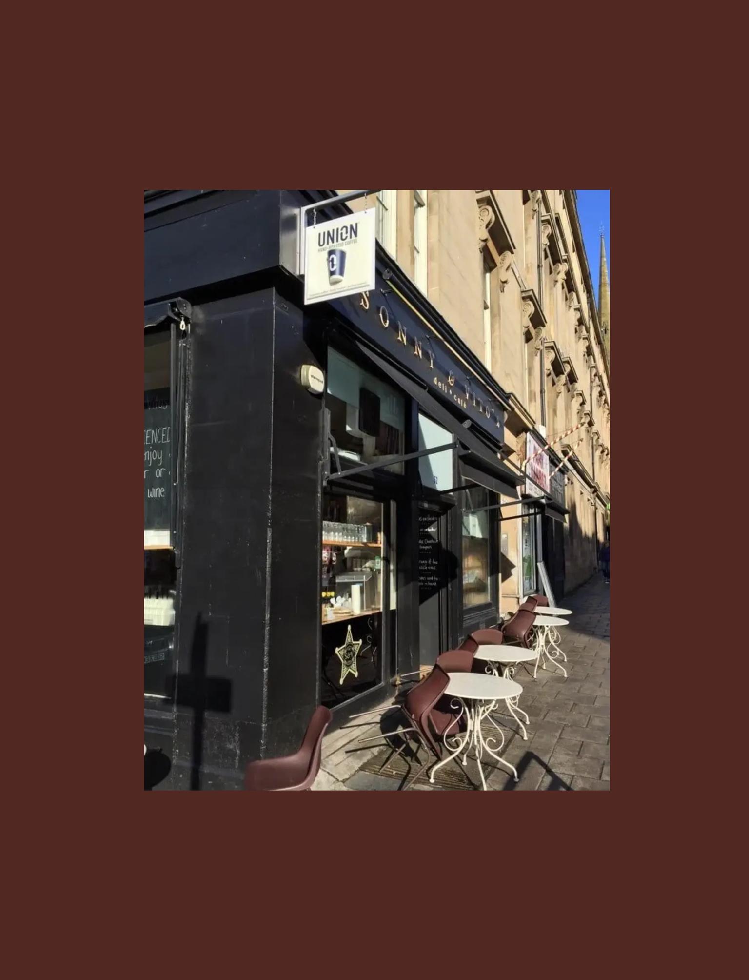 Exterior of a cafe on a corner, featuring outdoor seating with round tables, brown chairs, and a black facade under a sunny sky.