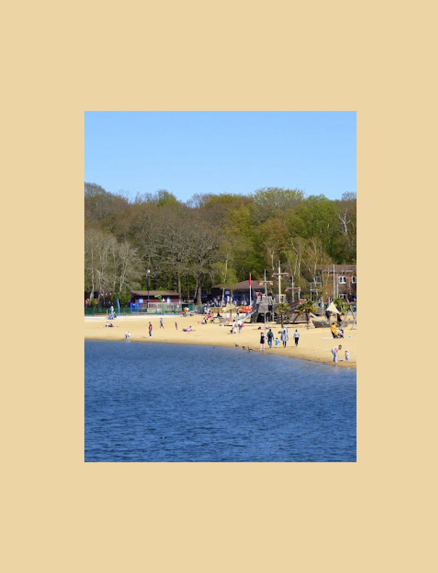 Scenic view of Ruislip Lido's sandy beach and playground bustling with families enjoying a sunny day by the water, surrounded by a leafy forest