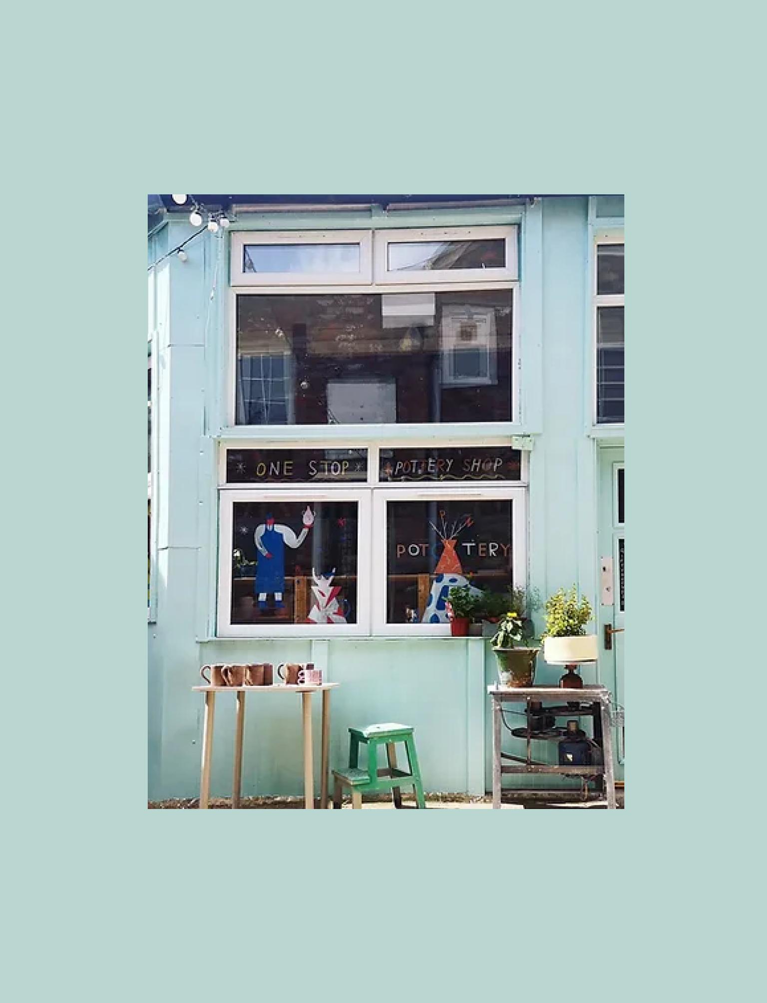 Spin Pottery Shop's pale blue facade features large windows displaying pottery and creative signs, inviting a warm, artistic vibe.
