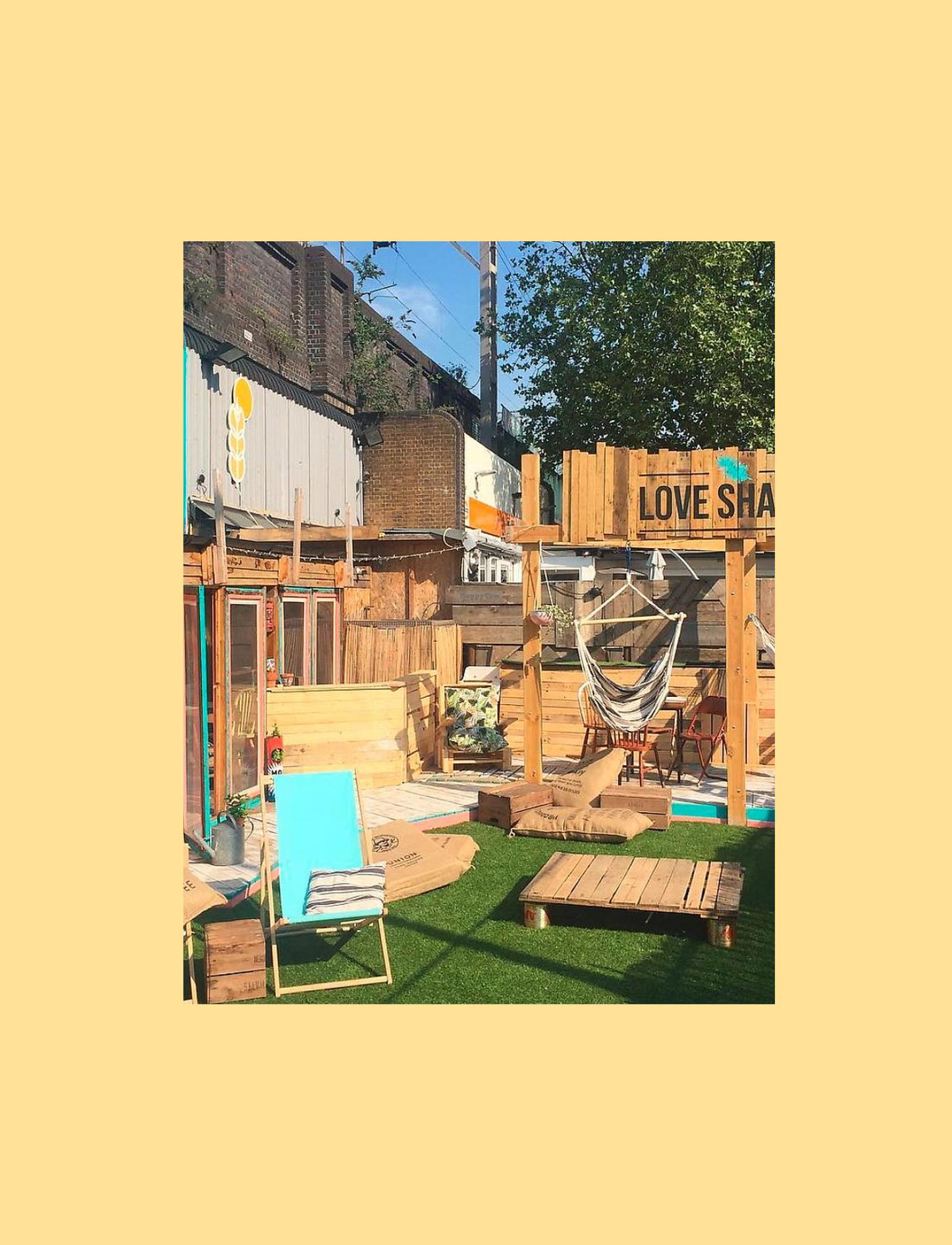 An outdoor restaurant space that shows multiple wooden decks and chairs for sitting. There is a small patch of astro turf at the front, upon which sits a blue deckchair.