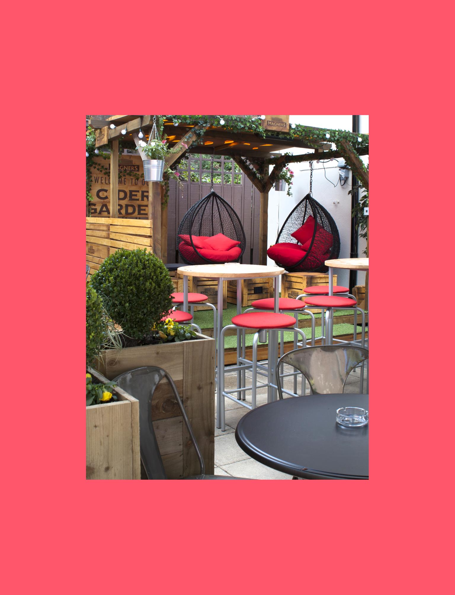 Cosy outdoor pub garden with vibrant red chairs, hanging swing seats, and lush greenery under a canopy.