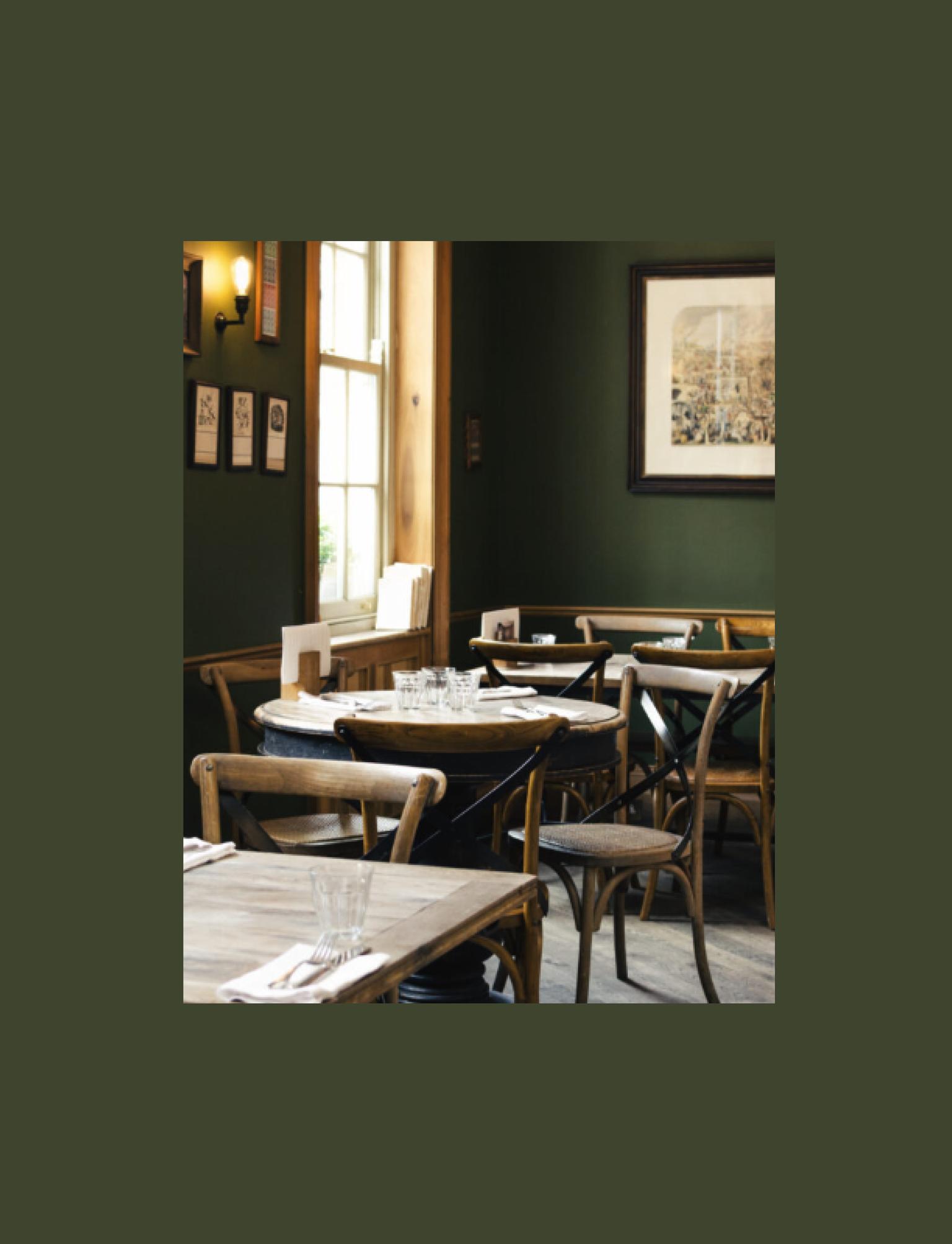 Intimate pub setting with dark green walls and wooden furniture. Natural light highlights the classic bentwood chairs and rustic tables set for dining. The walls are adorned with framed pictures and a large vintage-style painting.