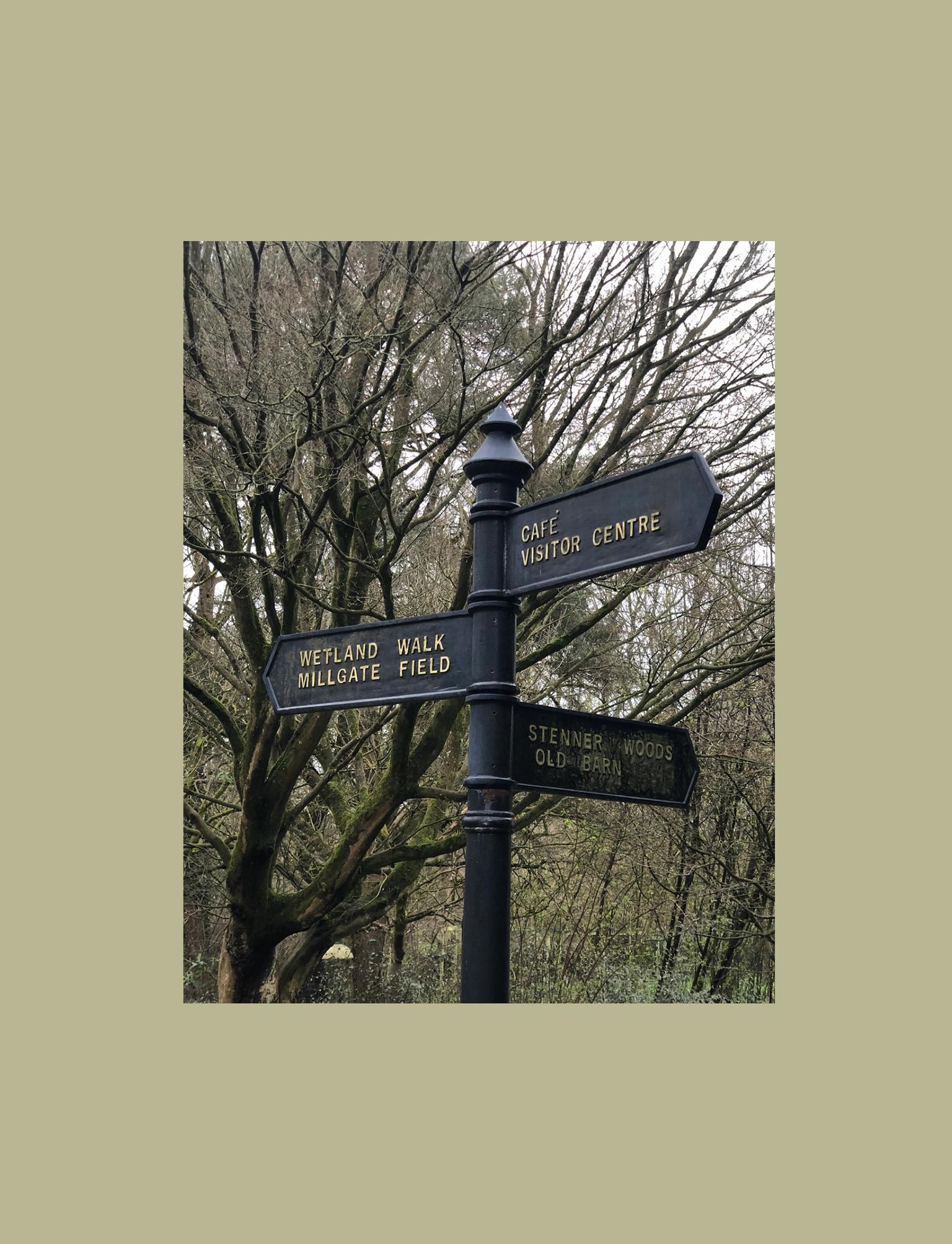 A directional signpost with arrows pointing to various locations, including "Wetland Walk", "Millgate Field","Cafe", "Visitor Centre", "Stenner Woods", and "Old Barn", set against a backdrop of leafless trees.
