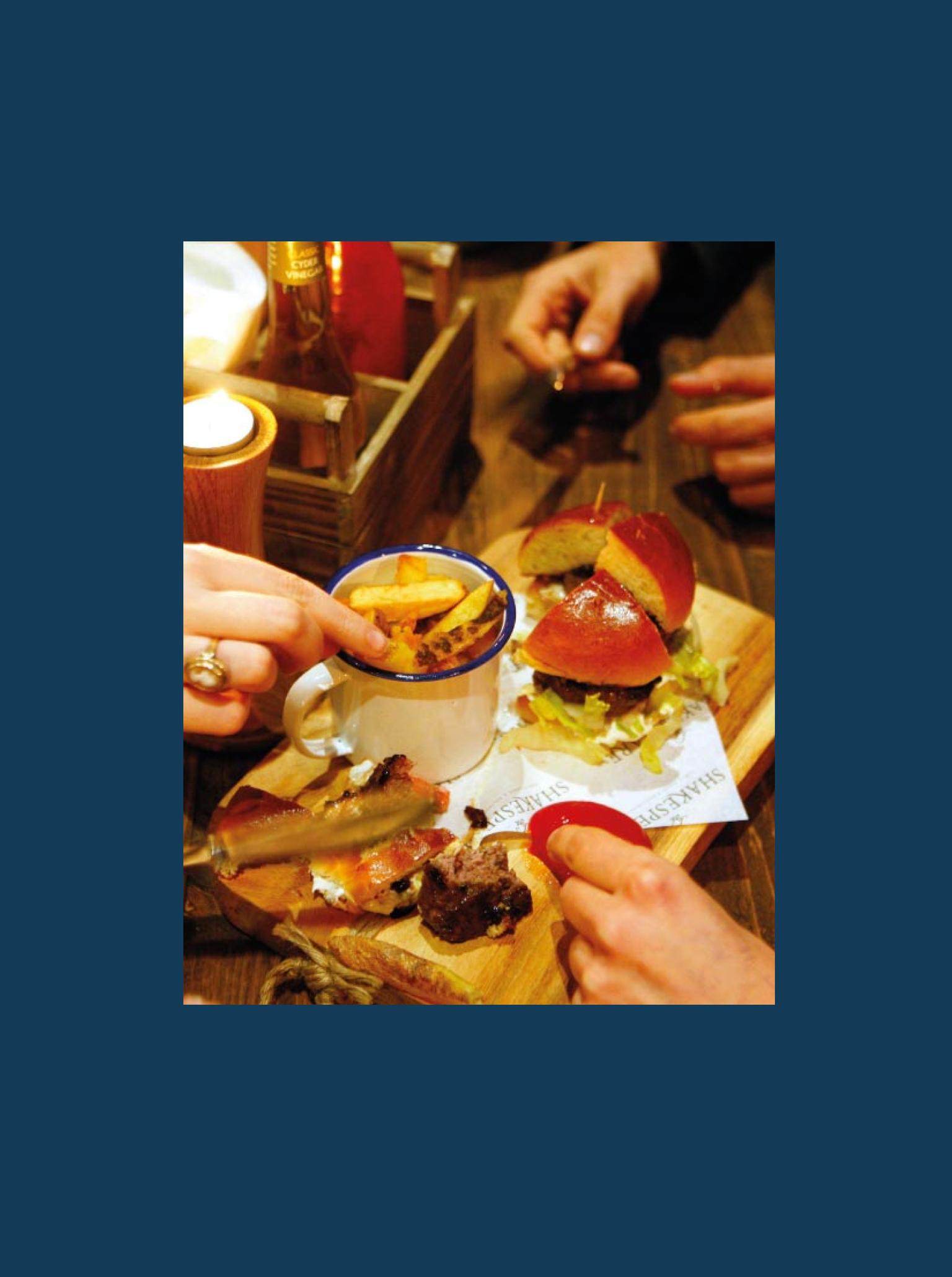 Four people's hands reaching towards a wooden board with a burger and chips on top of it.
