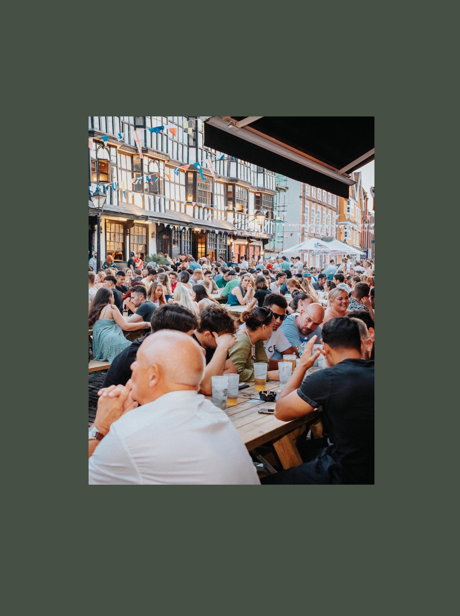 Lots of people sat drinking on pub benches in busy street