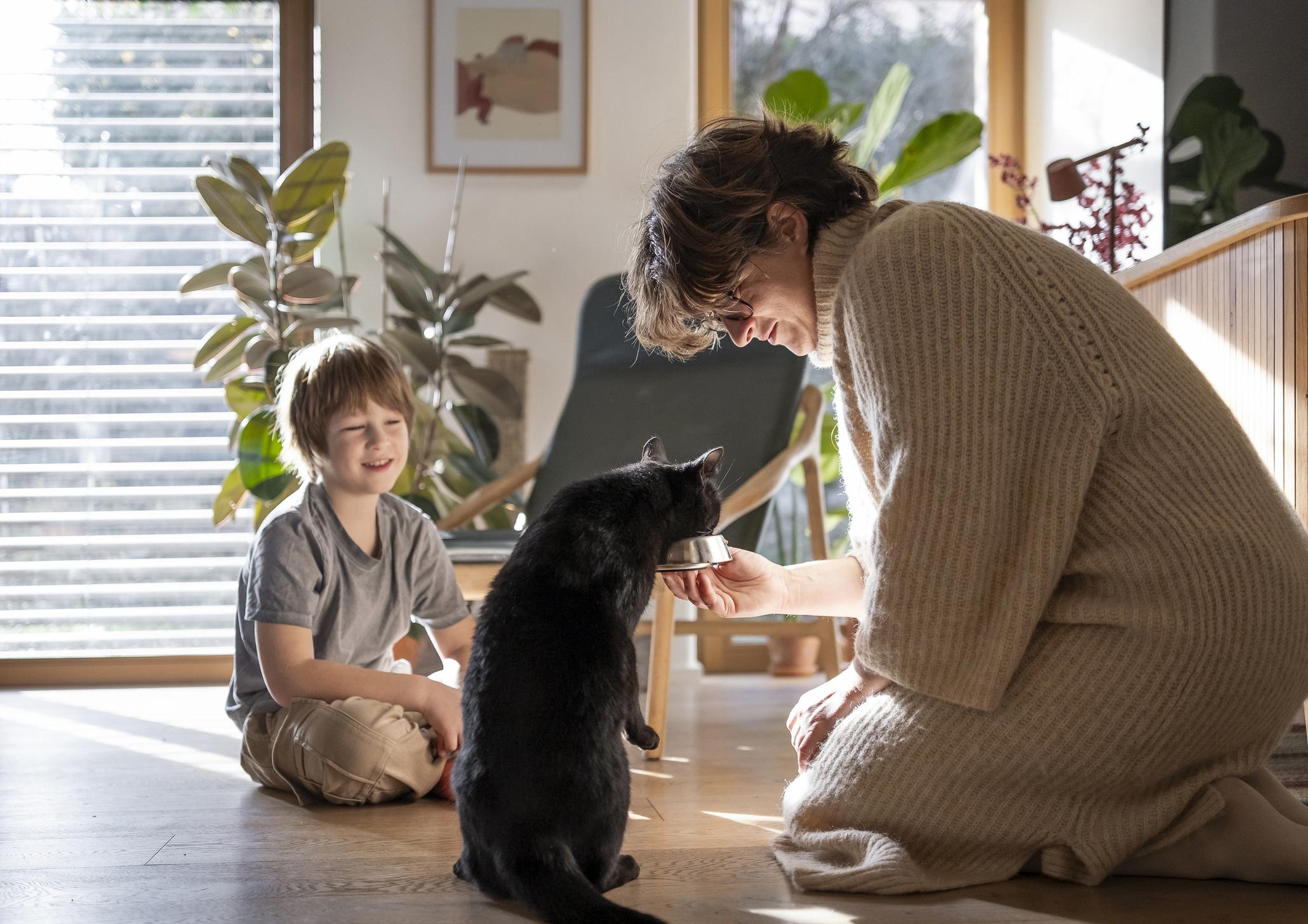 Woman in a cosy sweater gives food to a black cat while a child smiles, sitting on a sunlit wooden floor with plants in the background.