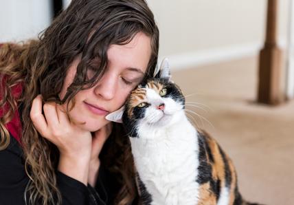 Woman with closed eyes gently leans her head against a calico cat, both exuding a sense of calm and affection.
