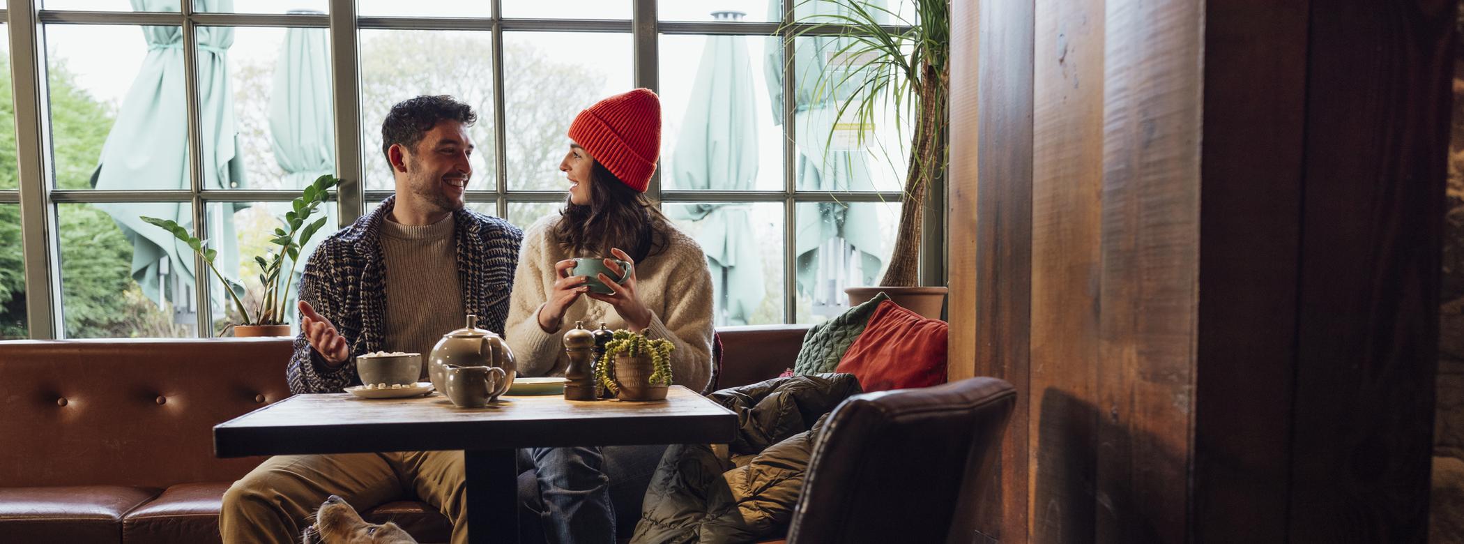 A couple enjoy a conversation in a pub or cafe with their Golden Retriever, who sits at their feet under the table and looks up at them