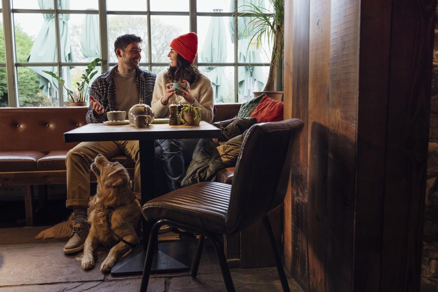A couple enjoy a conversation in a pub or cafe with their Golden Retriever, who sits at their feet under the table and looks up at them