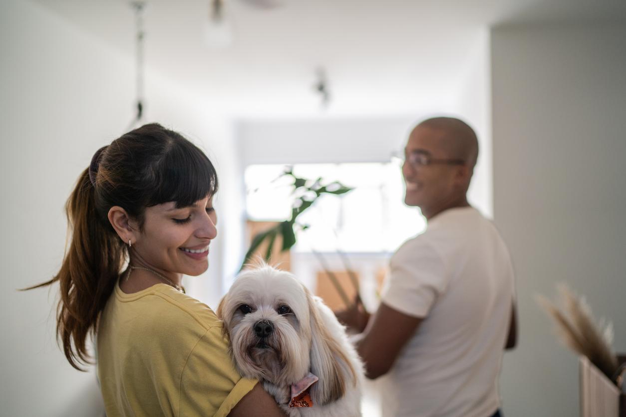A woman smiles while holding a fluffy dog. A man stands in the background holding a box in a bright room. It looks like the couple is moving into a new home.