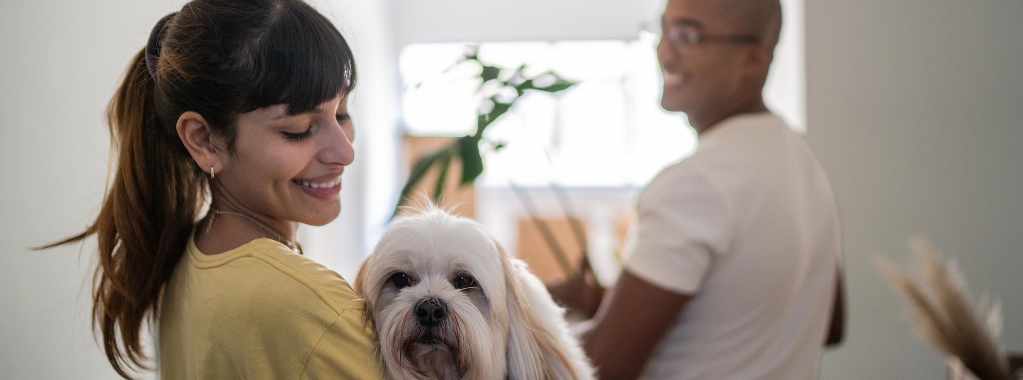 A woman smiles while holding a fluffy dog. A man stands in the background holding a box in a bright room. It looks like the couple is moving into a new home.