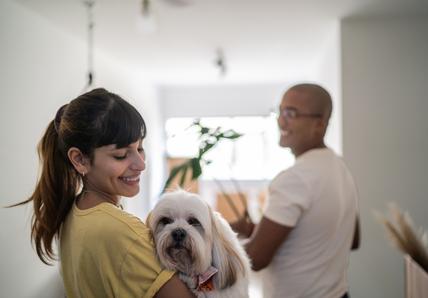 A woman smiles while holding a fluffy dog. A man stands in the background holding a box in a bright room. It looks like the couple is moving into a new home.