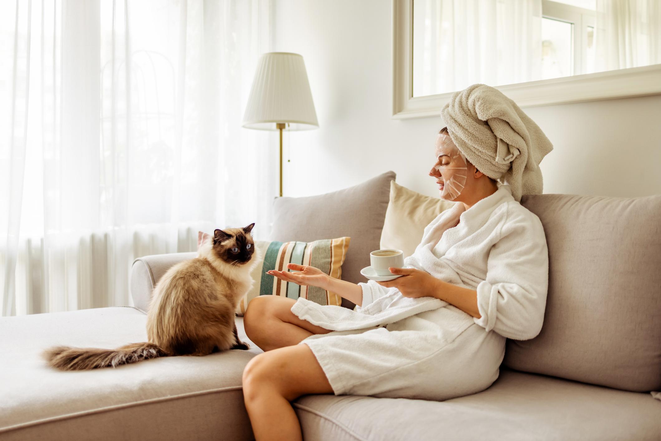 A woman in a dressing gown, towel, face mask and a cup of tea relaxes with their cat
