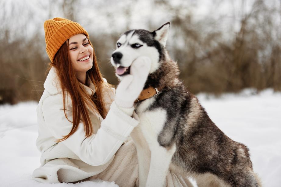 A woman with long red hair wearing an orange hat pets a husky dog outside in the snow