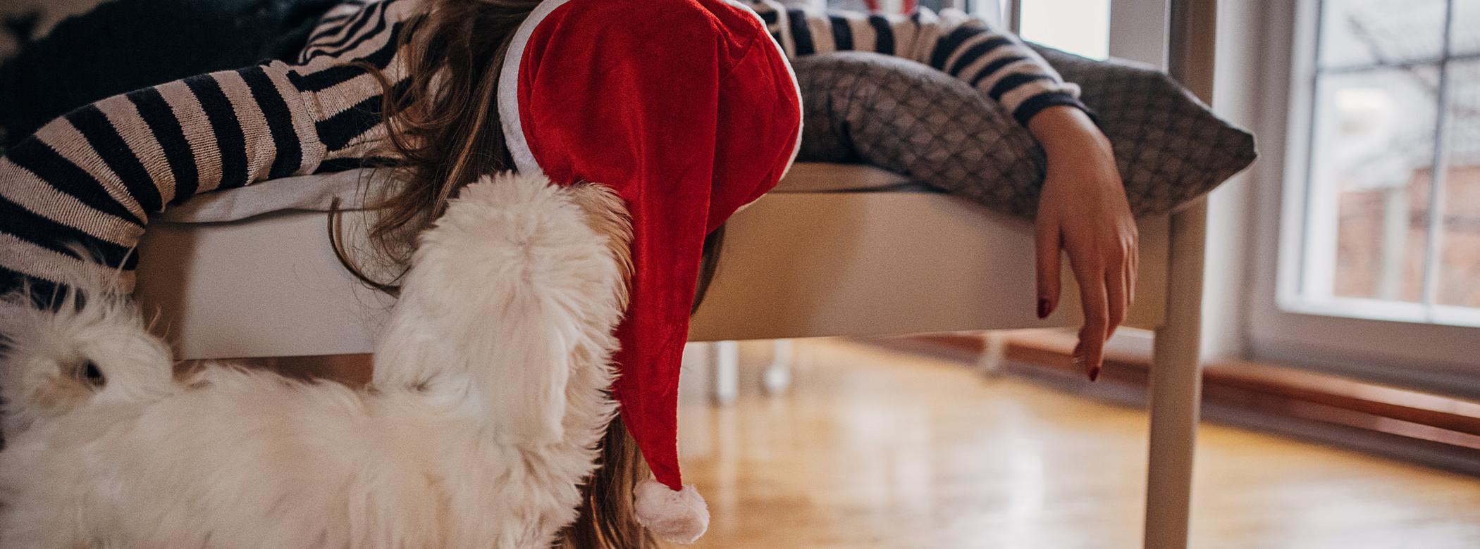A woman in a Santa Hat lies down hungover, her dog is sniffing her to see if she&#39;s okay