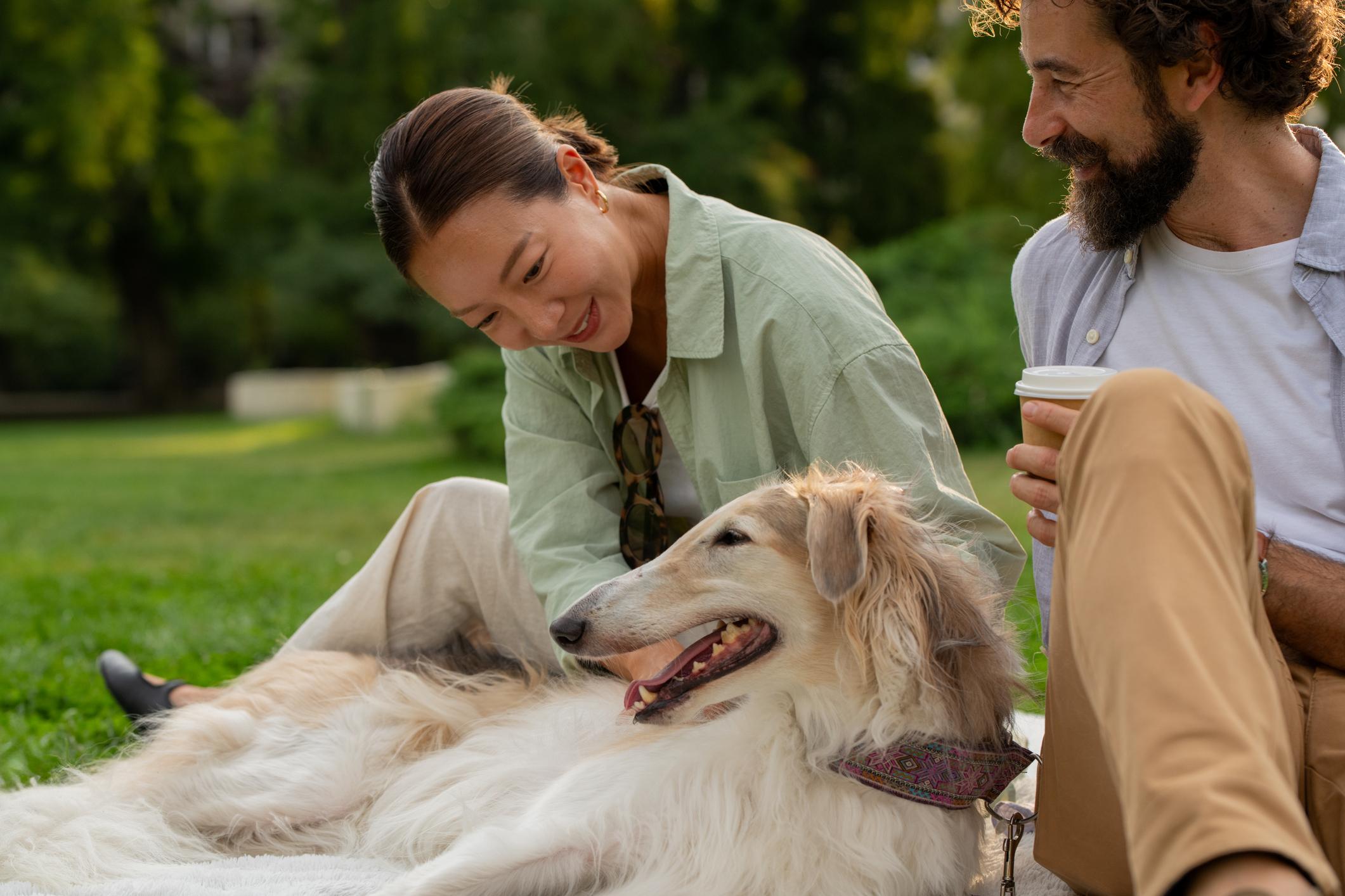 A joyful couple sits on grass, petting a relaxed Borzoi dog. The woman smiles at the dog; the man holds a coffee cup. Lush greenery is in the background.
