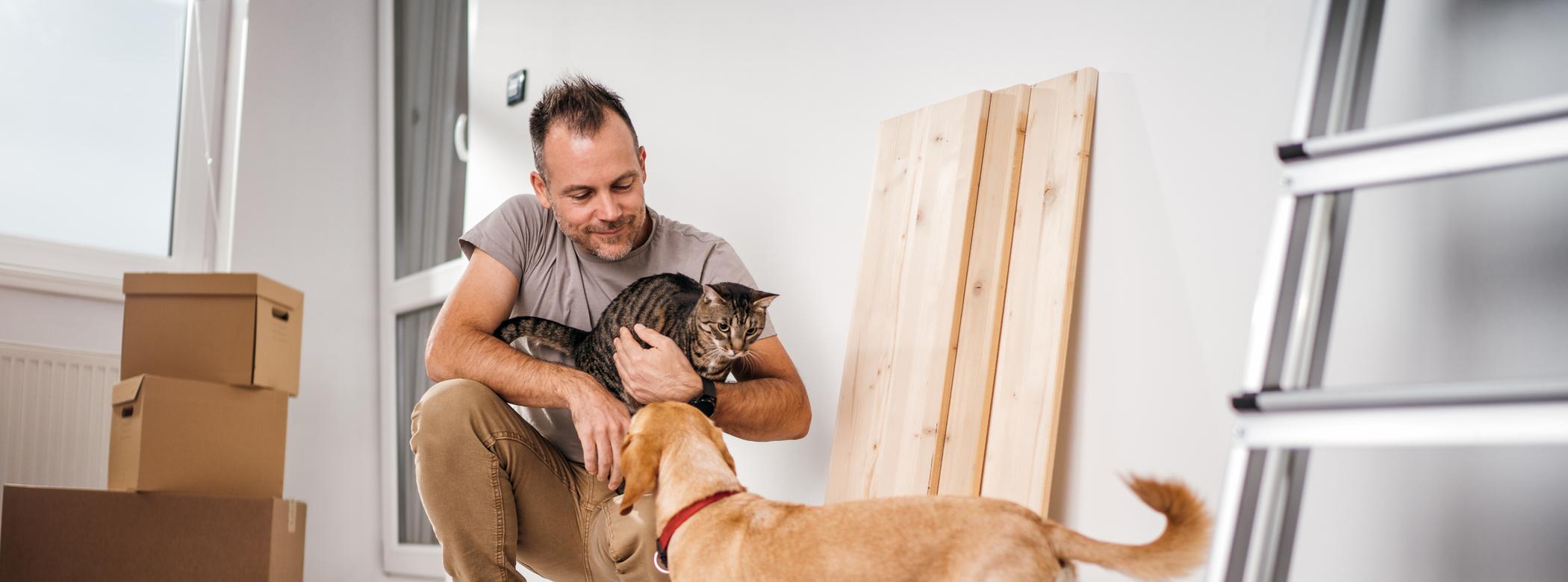 A man holds his cat and looks at his dog while he's in the middle of renovating his home, he's crouched down and there are home upgrade materials around him