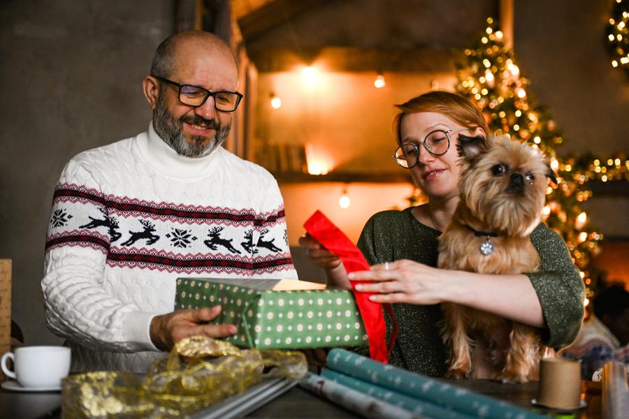 A man and a woman, likely a couple, wrap gifts while a dog is sat on the woman's lap